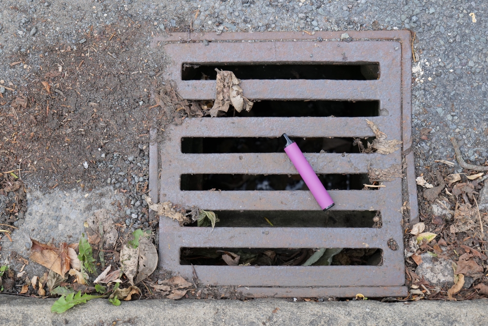 A purple e-cigarette vape has been discarded and left lying on a metal water drain cover.