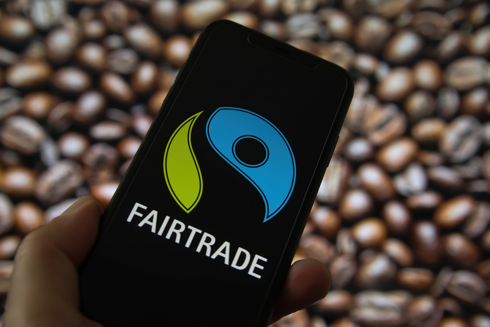 Closeup of mobile phone screen with logo lettering of fairtrade label seal, blurred coffee beans background (focus on left lower part of blue logo)